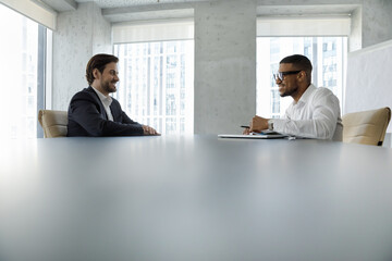 Side view African and Caucasian businessmen, applicant and HR manager sit at desk in modern office board room at formal meeting or negotiations. Headhunter and candidate during job interview concept