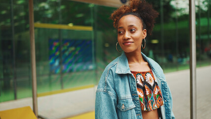 Cute African girl with ponytail, wearing denim jacket, in crop top with national pattern, standing at the bus stop and smiling