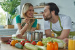 © gstockstudio - Happy woman giving her husband to taste a meal while both standing at the kitchen