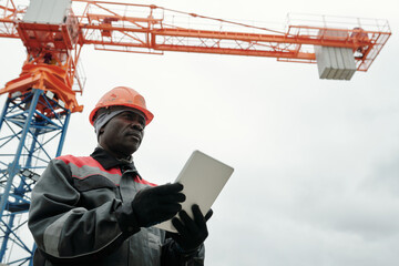 Poster - Serious black man in workwear, and hardhat using tablet while standing at construction site against working tower crane and looking forwards