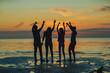 © Comeback Images - Silhouettes of four diverse young friends dancing with beer bottles in raised hands by sea at orange sunset during beach party, backlit back view