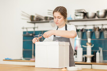 Young Female Bakery Chef Packaging Freshly Made Cake Into Gift Box, Tying It And Cutting String With Scissors Standing At Table In Restaurant Kitchen