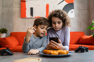 Wall Mural - Siblings brother and sister boy and girl or children friends using mobile phone smartphone at home in room to watch video make a call or play online games leisure family concept real people copy space