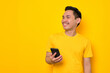© Sewupari Studio - Smiling young Asian man in casual t-shirt holding mobile phone and looking at aside isolated on yellow background. People lifestyle concept