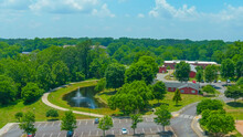 An Aerial Shot Of A Gorgeous Summer Landscape In The Park With A Pond With A Water Fountain Surrounded By Lush Green Trees, Grass And Plants With Red Buildings At Logan Farm Park In Acworth Georgia