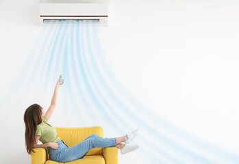 Poster - Young woman switching on air conditioner while sitting in armchair near white wall