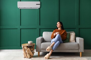 Beautiful young woman with cup of tea resting on sofa in room with operating air conditioner