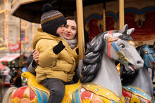 Woman Supporting Boy Sitting On Carousel Horse In Christmas Market