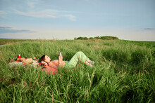 Young Woman Reading Book Lying On Grass On Field At Sunset
