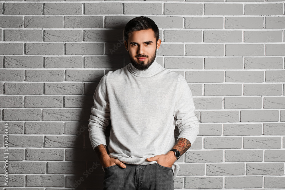 Young bearded man keeping hands in pockets near grey brick wall