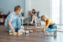Siblings Playing With Toy Train Set While Mother Using Smart Phone Sitting On Armchair At Home