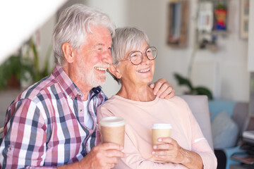 Wall Mural - Amused senior couple at home hugging while holding two cups of coffee. Strong relationship for two Caucasian seniors who are comfortable together