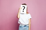 Fototapeta  - Pretty woman covering face using a white paper sheet with drawn question mark, like a mask, for hiding her identity on pink background