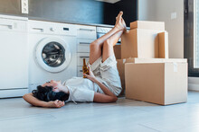Happy Woman Holding Beer Bottle With Feet Up On Cardboard Box Lying By Washing Machine In Utility Room