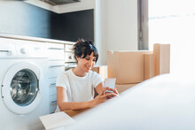 Smiling Woman Using Smart Phone Sitting With Cardboard Boxes By Washing Machine In Utility Room
