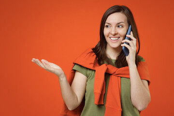 Wall Mural - Young smiling happy woman 20s wear khaki t-shirt tied sweater on shoulders look aside hold in hand use mobile cell phone isolated on plain orange background studio portrait. People lifestyle concept.