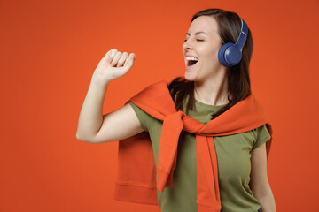 Wall Mural - Young happy fun woman 20s in khaki t-shirt tied sweater on shoulders headphones listen to music sing song in microphone isolated on plain orange background studio portrait. People lifestyle concept.