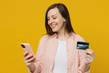 Wall Mural - Young happy woman she 30s wears striped shirt white t-shirt using mobile cell phone hold credit bank card doing online shopping order delivery booking tour isolated on plain yellow background studio.