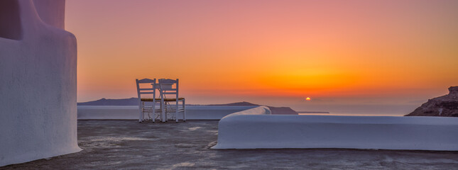 White architecture in romantic sunset light on Santorini island, Greece. Couple chairs on caldera terrace with sea view, beautiful colorful skyline sky view. Honeymoon panoramic travel background