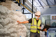© เลิศลักษณ์ ทิพชัย - A male worker using a tablet computer working in an industrial chemical or parcel factory wears a white helmet in an industrial warehouse. The background is a warehouse and goods.
