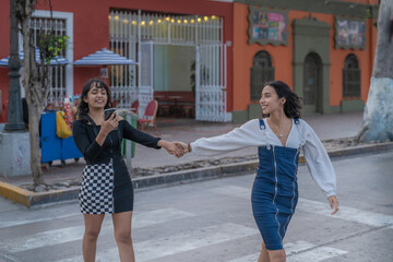 Girl looking at the mobile crossing the street while holding hands with a friend