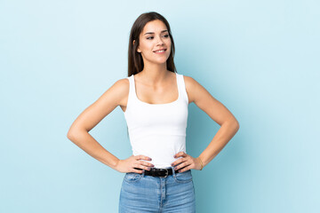 Sticker - Young caucasian woman isolated on blue background posing with arms at hip and smiling