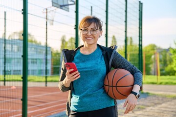 Wall Mural - Active mature woman with basketball ball and smartphone, outdoor