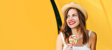 Young Ukrainian Woman With Red Lipstick In A Straw Hat And A White Dress With A Lollipop Smiles On A Yellow Background, Copy Space. Web Banner