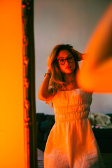 Wall Mural - Young attractive woman looking on herself in the mirror in the evening in orange light.