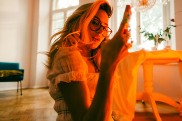 Wall Mural - Portrait of attractive caucasian woman fashion model in eyeglasses and white dress posing while sitting on a floor in living room with vintage decor.