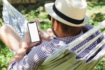 Man in hat using his smartphone while lying in hammock and relaxing.