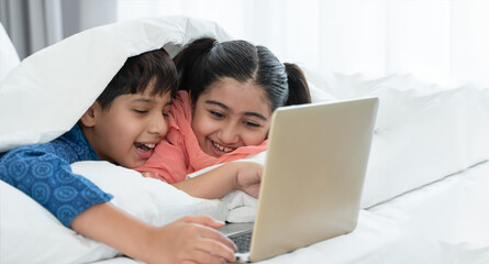 Wall Mural - Two happy Indian brother and sister in traditional clothing lying on bed under blanket smiling, using laptop, having fun together at home. Education, Siblings relationship concept