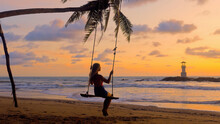 Young Woman Swinging Sitting On Rope Swing Suspended From Palm Tree At Sea Beach In Sunset Lights. Girl Silhouette Enjoy Outdoors Freedom, Summer Relax. Travel, Tourist, Holidays, Vacations Concept.
