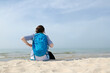 Dziewczyna, kobieta z plecakiem siedzi na piaszczystej morskiej plaży. Wpatruje się w horyzont. Lato, pastelowe kolory, przydymione błękitne niebo