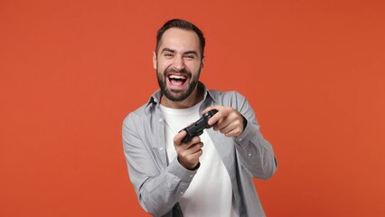 Wall Mural - Blithesome charismatic gambling overjoyed excited young brunet man 20s years old wears blue shirt hold in hand play pc game with joystick console isolated on plain orange background studio portrait