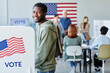 © Seventyfour - Side view portrait of smiling black man voting in booth on election day and looking at camera, copy space