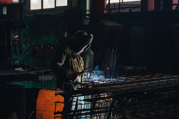 Wall Mural - Factory worker in protective mask welding iron armature reinforcement for concrete