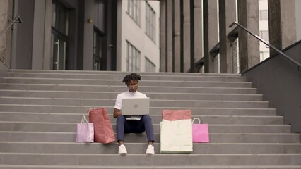 Wall Mural - City shopping concept. Outdoor urban lifestyle portrait of diverse young African American woman, sitting on the steps of the mall with a laptop and colorful shopping bags while shopping online.