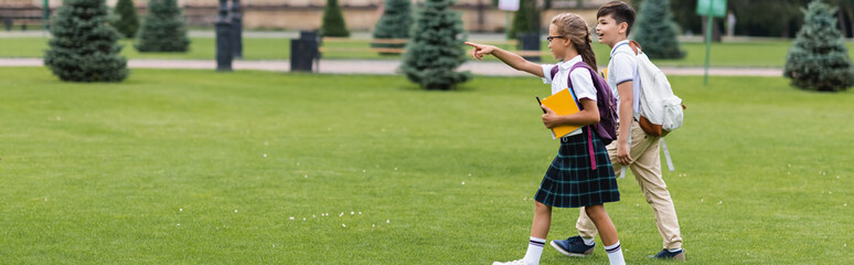 Wall Mural - Side view of schoolgirl pointing with finger near asian friend while walking on lawn, banner.