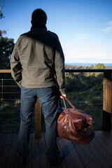 caucasian man behind holding brown leather travel overnight bag blue pants green jacket outdoors view mountains balcony