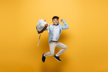 Wall Mural - Cheerful asian schoolkid holding backpack and jumping on yellow background.