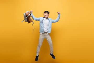 Wall Mural - Excited asian schoolboy holding backpack and showing yes gesture while jumping on yellow.