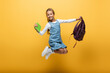 Smiling schoolkid with backpack and book jumping on yellow background.
