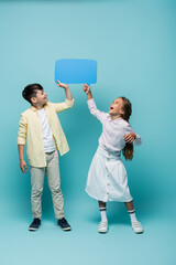 Wall Mural - Cheerful interracial schoolchildren holding speech bubble on blue background.