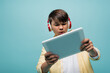 Focused asian schoolkid in headphones using digital tablet isolated on blue.