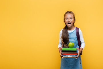 Wall Mural - Cheerful pupil holding books and apple isolated on yellow.