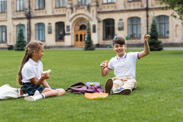 Wall Mural - Asian schoolboy holding sandwich and pointing with finger near backpack and classmate in park.