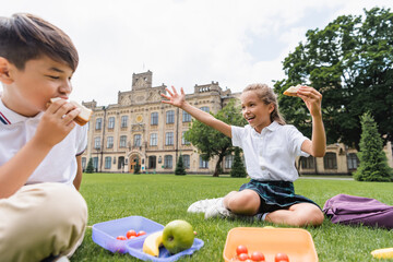 Wall Mural - Schoolgirl holding sandwich and waving hand near lunchboxes and blurred asian friend on grass.