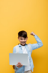 Wall Mural - Excited asian schoolkid showing yes gesture and holding laptop isolated on yellow.