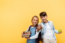 Smiling Multiethnic Pupils With Books And Apple Using Mobile Phone Isolated On Yellow.